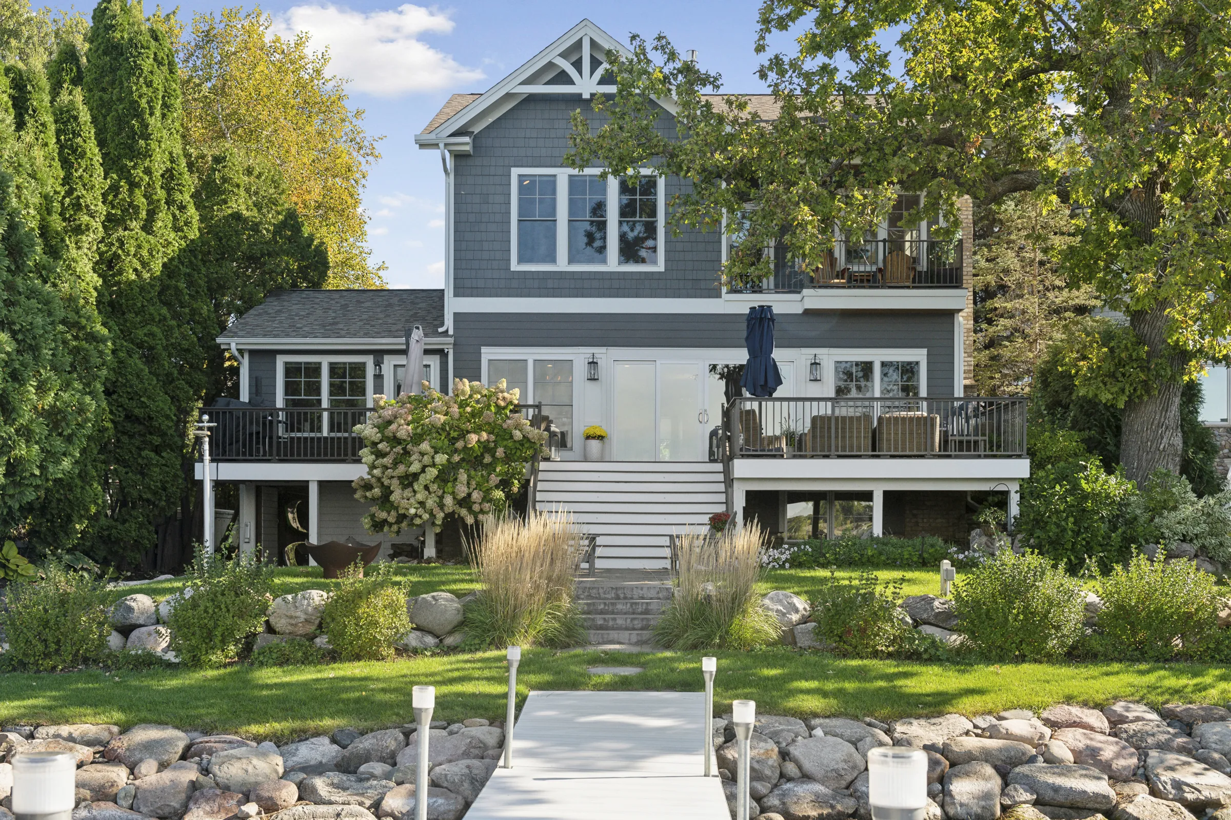 Photo of back of remodeled house with gray siding and white trim and a newly landscaped lawn with shrubs and a rock wall